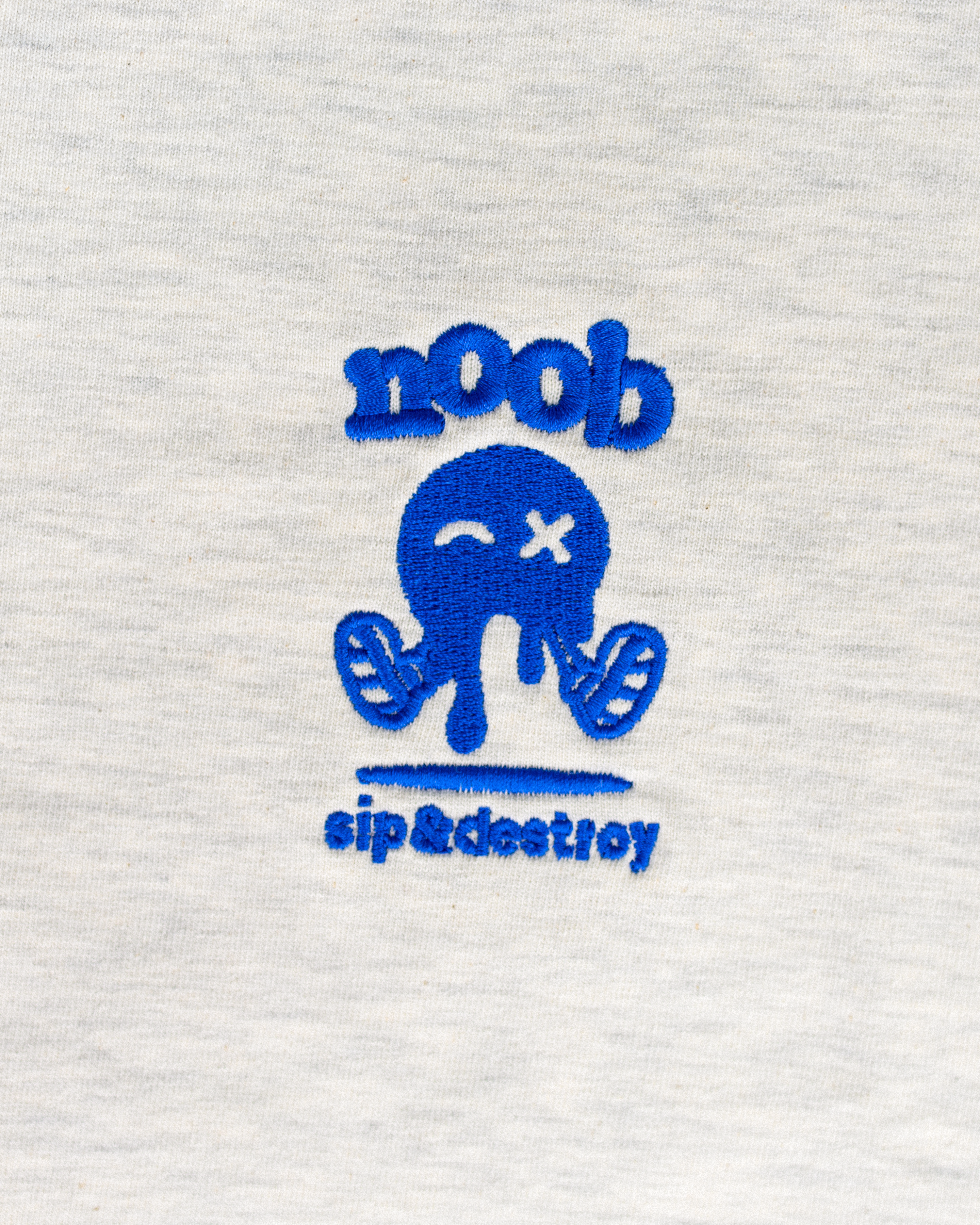 NOOB Spanky Logo embroidered in blue on the font of a heather oatmeal t-shirt showing close up of stitching.